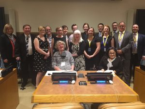 Fifteen people stand and two people sit in front of a large wooden desk in a conference room