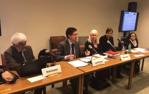 Five people sit behind wooden desks with various papers and microphones in a conference room. A large nameplate is in on the desks in front of each person.